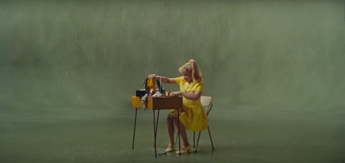 A screenshot from the music video for the song What Was I Made For features Eilish clad in yellow with a high pony, unpacking clothes for her Barbies. 